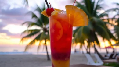 A vibrant, colorful tropical drink served in a tall glass filled with layers of sunset hues — deep oranges, pinks, and purples — garnished with fresh fruit slices like pineapple and cherries, surrounded by ice cubes, with a backdrop of a serene beach at sunset, palm trees swaying gently in the breeze.